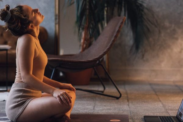 A woman doing yoga on a mat indoors, facing a laptop, in a serene setting.