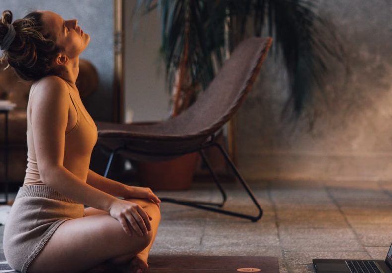 A woman doing yoga on a mat indoors, facing a laptop, in a serene setting.